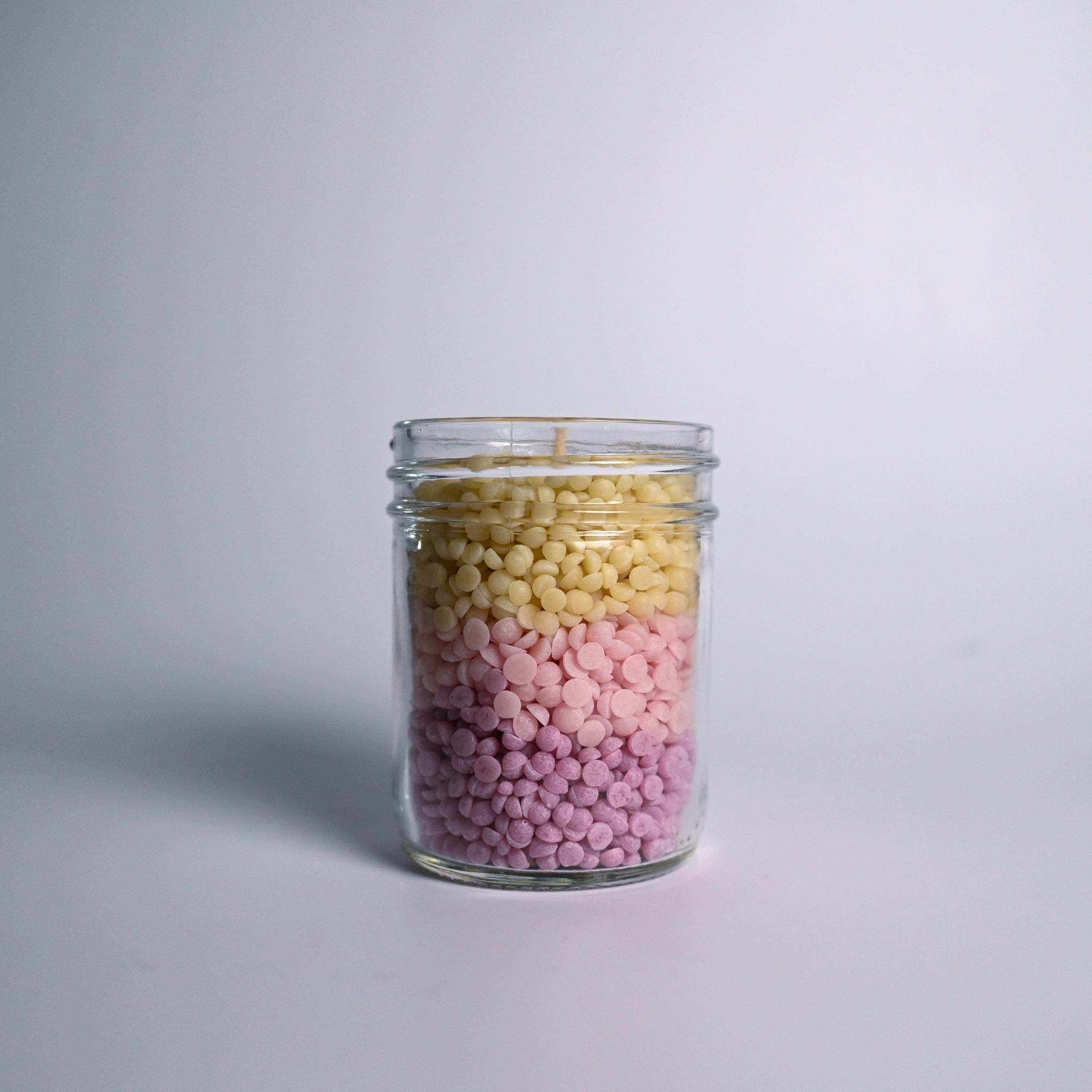 Glass jar filled with purple, pink and yellow wax beads on a light gray background