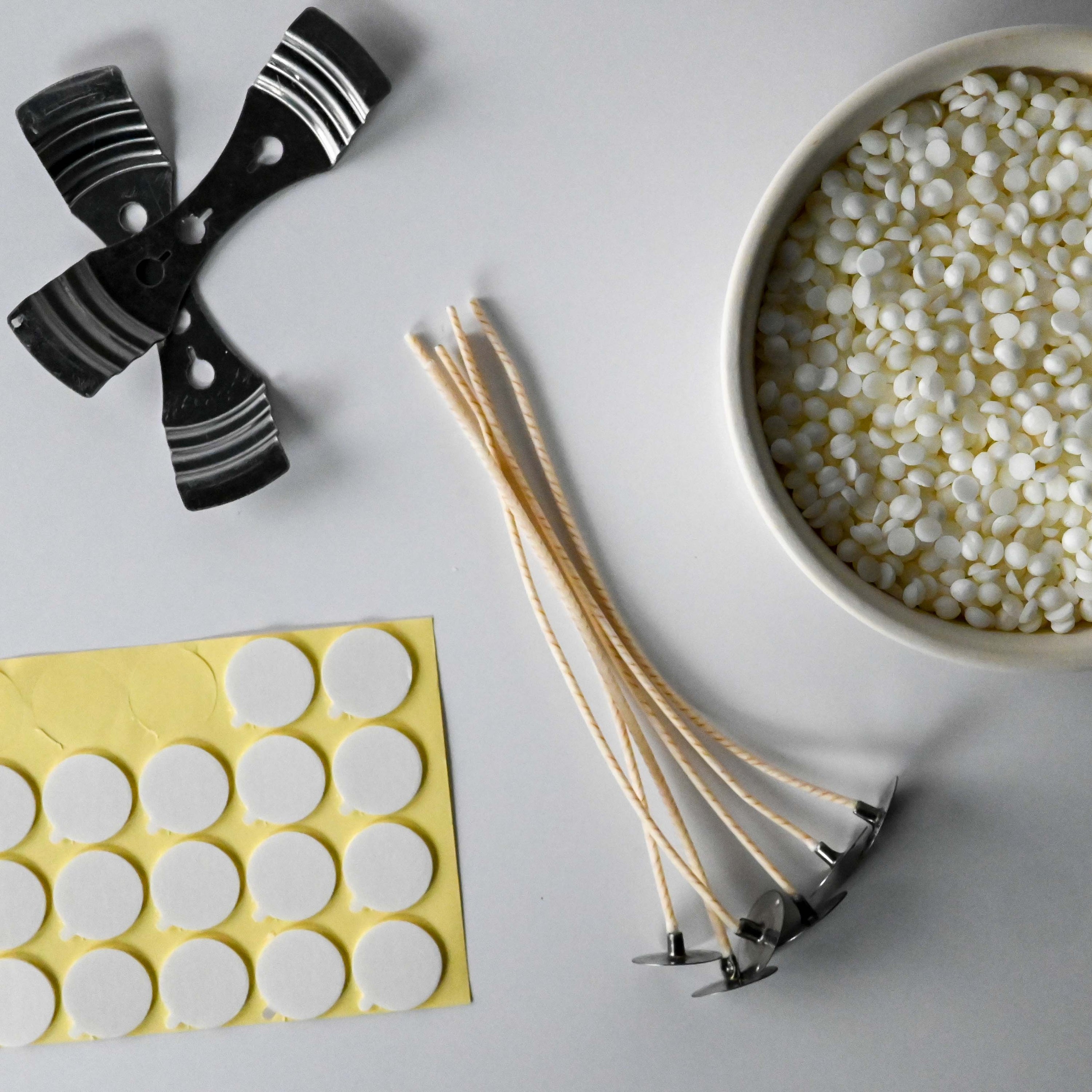 Candle-making supplies including wick holders, wax beads, wicks, and adhesive rounds on a white background