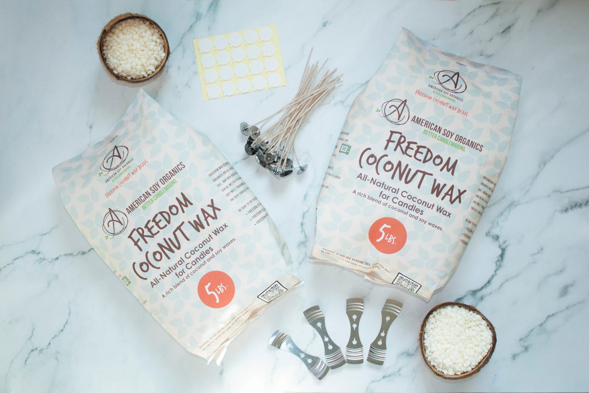 Two 5 lb. packages of Freedom Coconut Wax with wicks, adhesive rounds, wick holders and a glass containers with wax beads, displayed on a marble surface.