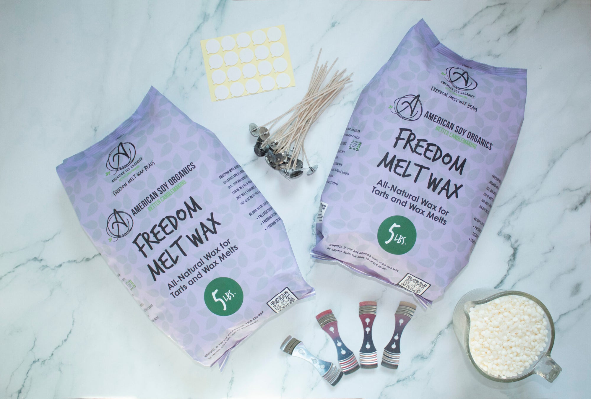 Two 5 lb. packages of Freedom Melt Wax with wicks, adhesive rounds, wick holders and a glass container with wax beads, displayed on a marble surface.
