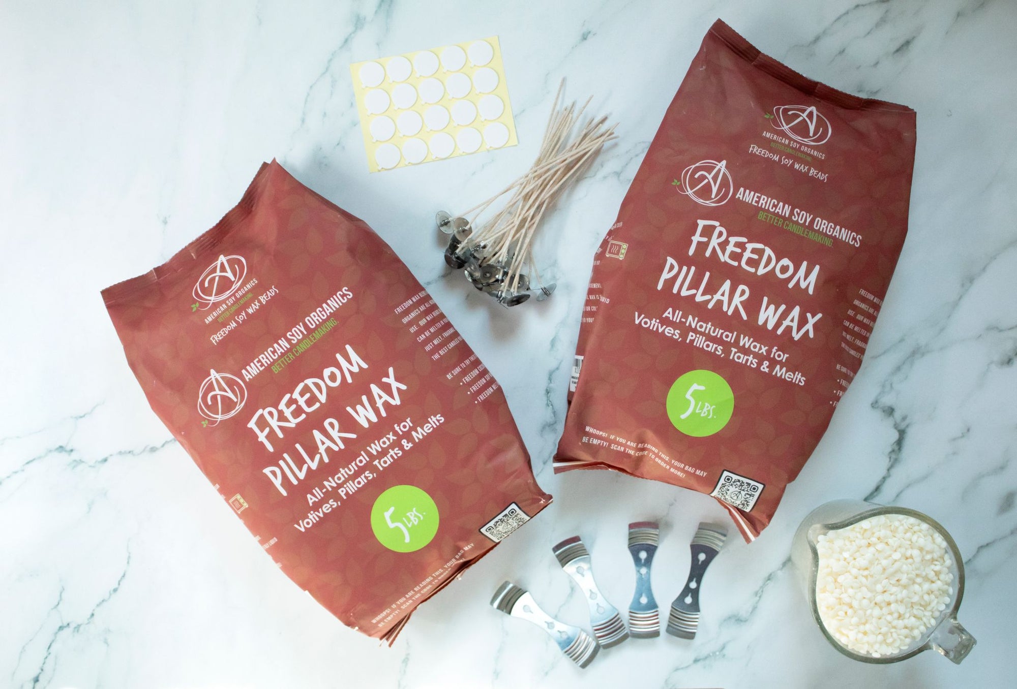 Two 5 lb. packages of Freedom Pillar Wax with wicks, adhesive rounds, wick holders and a glass container with wax beads, displayed on a marble surface.