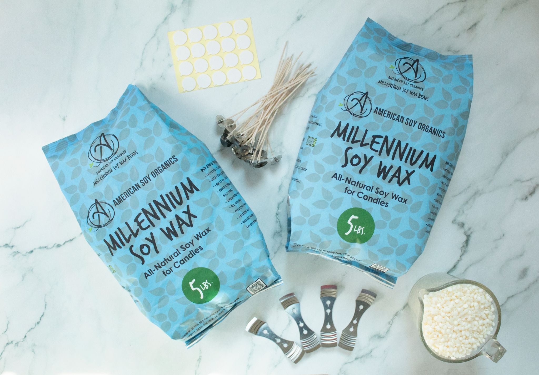 Two 5 lb. packages of Millennium Soy Wax with wicks, adhesive rounds, wick holders and a glass container with wax beads, displayed on a marble surface.