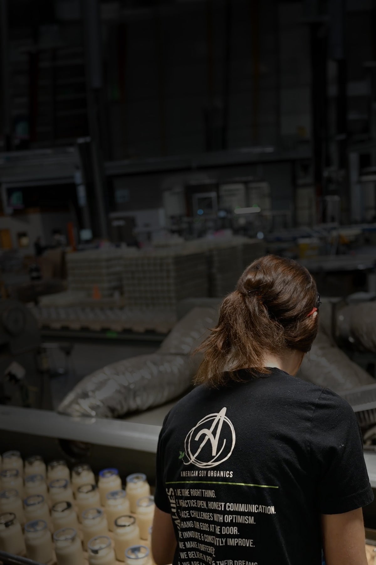 Person wearing a black t-shirt with a logo and text, standing in a warehouse setting.