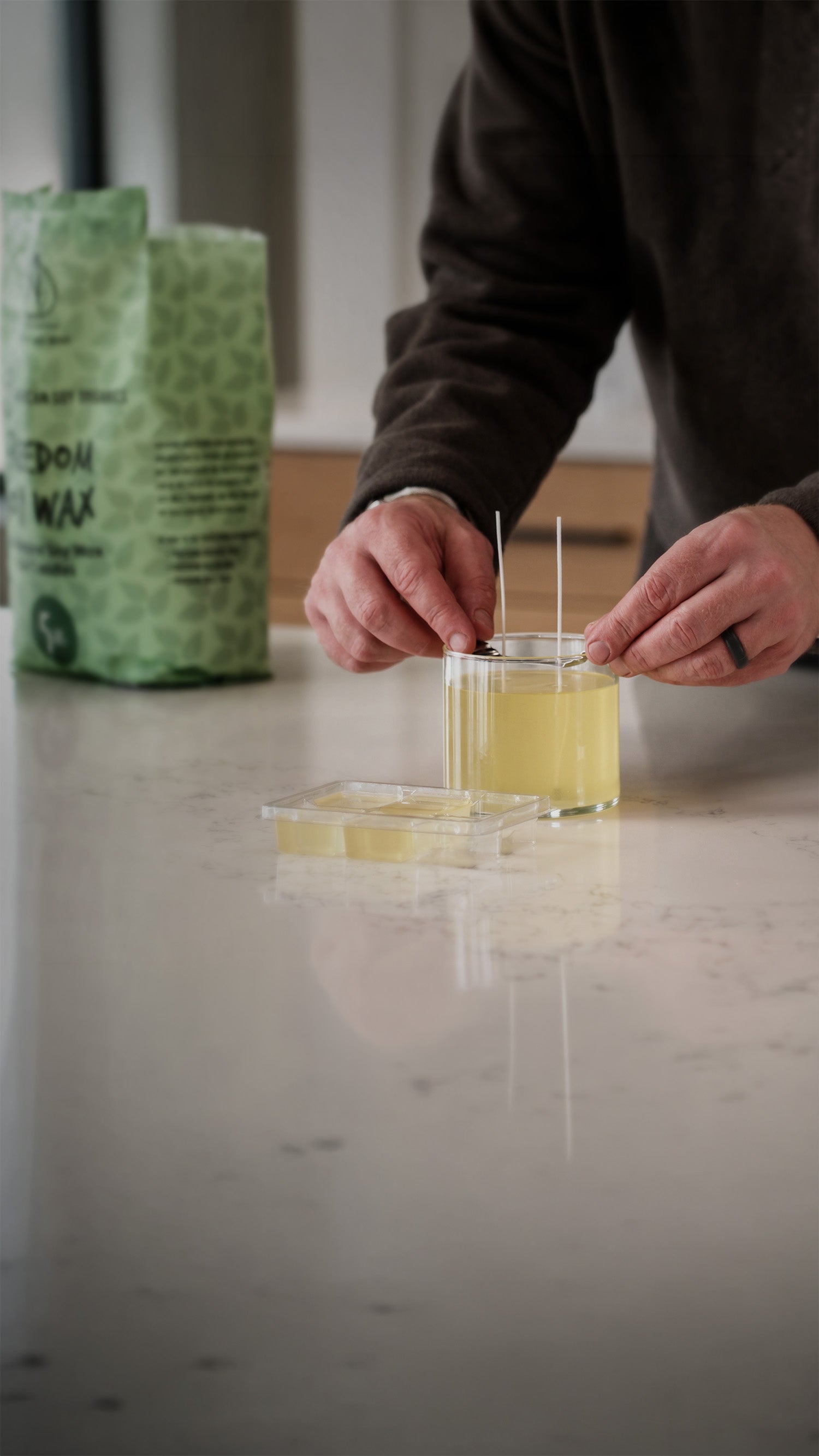 Person pouring liquid wax into a small container on a marble surface with a green package in the background.