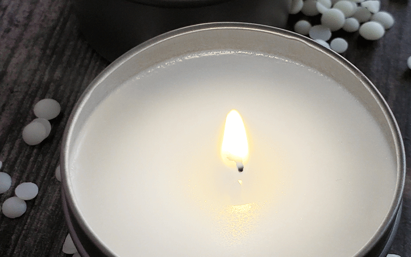 Close-up of a candle in a small tin vessel, set against a distressed wooden background, with a few white wax beads nearby