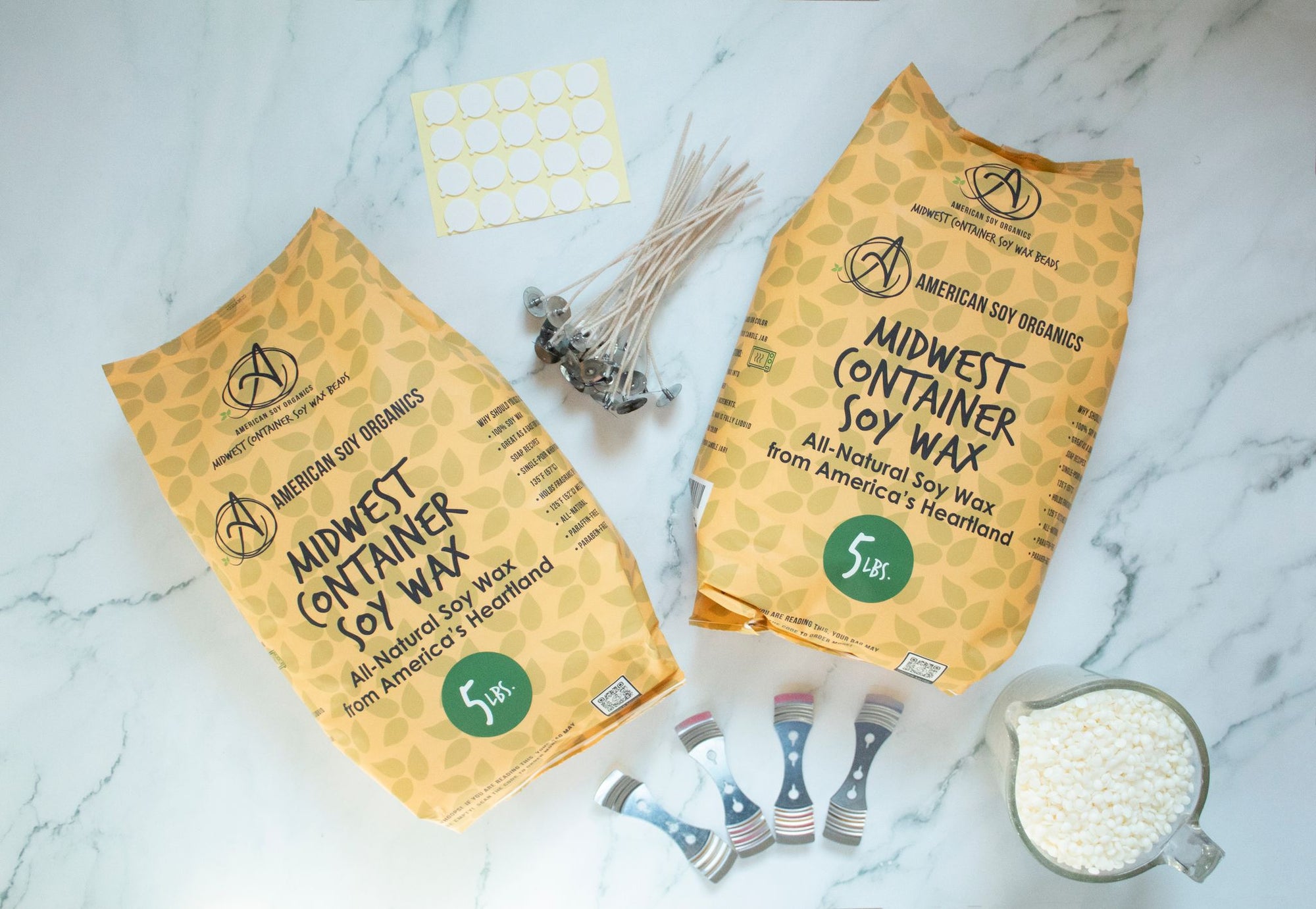 Two 5 lb. packages of Midwest Container Soy Wax with wicks, adhesive rounds, wick holders and a glass container with wax beads, displayed on a marble surface.